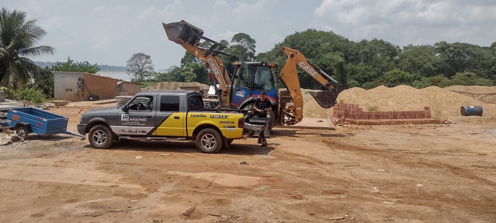 caminhonete com adesivos da LS Máquinas e retroescavadeira Case em pátio de terra com pilas de areia e tijolos