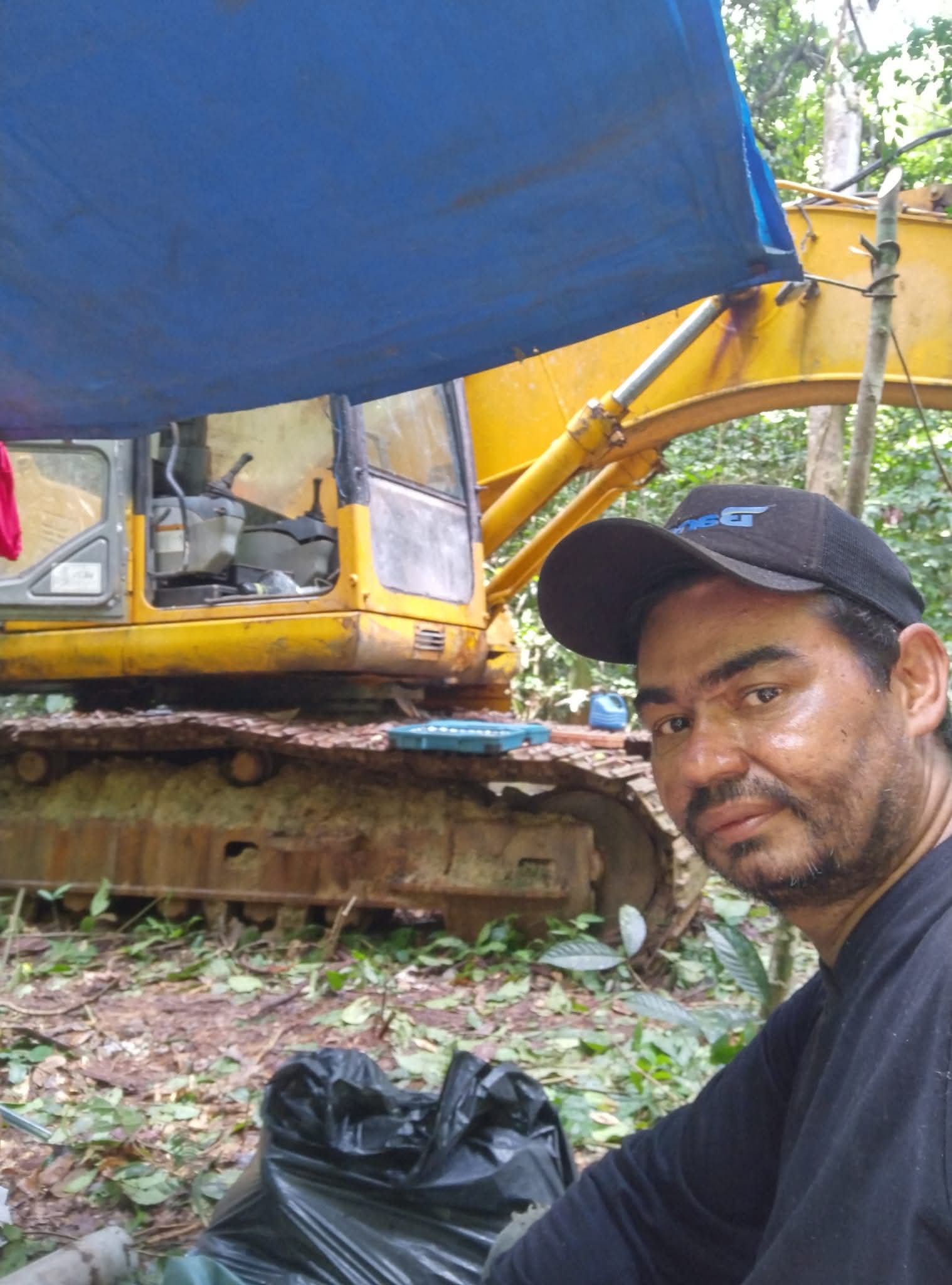 homem de boné preto e camiseta escura faz selfie em frente a uma escavadeira amarela no meio da mata, com lona azul em c