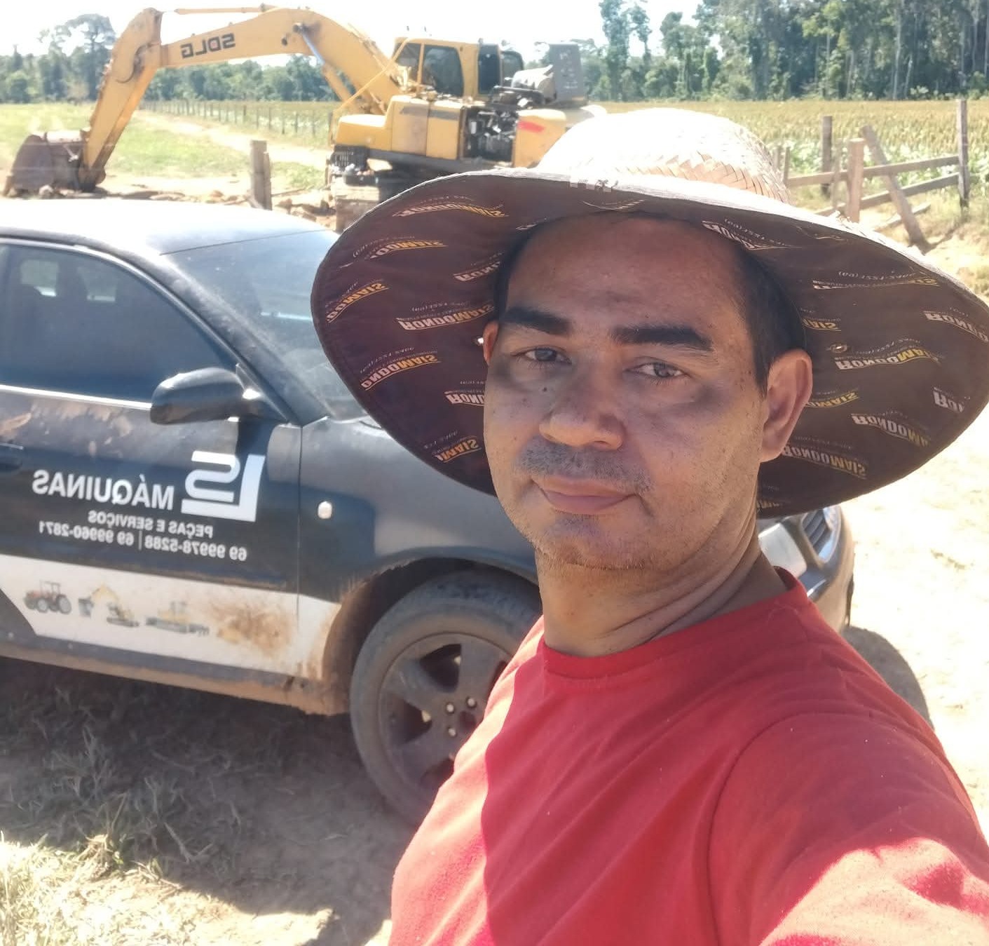 homem de chapéu e camisa vermelha tira selfie ao lado de carro da empresa LS Máquinas Peças e Serviços, com es